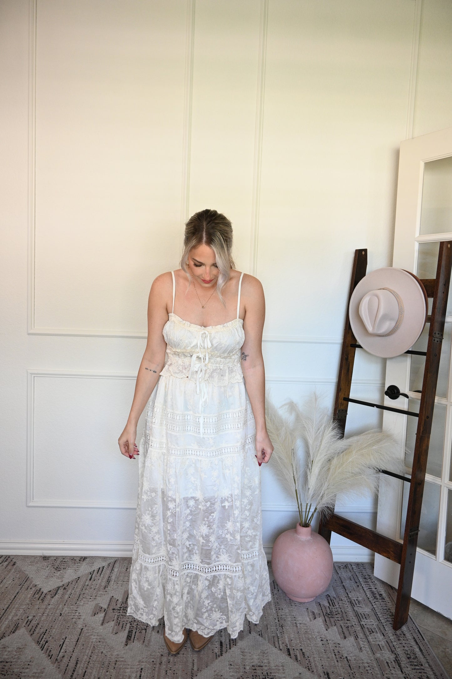 Woman in a ivory tank top and lace skirt set standing next to a hat on a stand in a room with a textured wall.