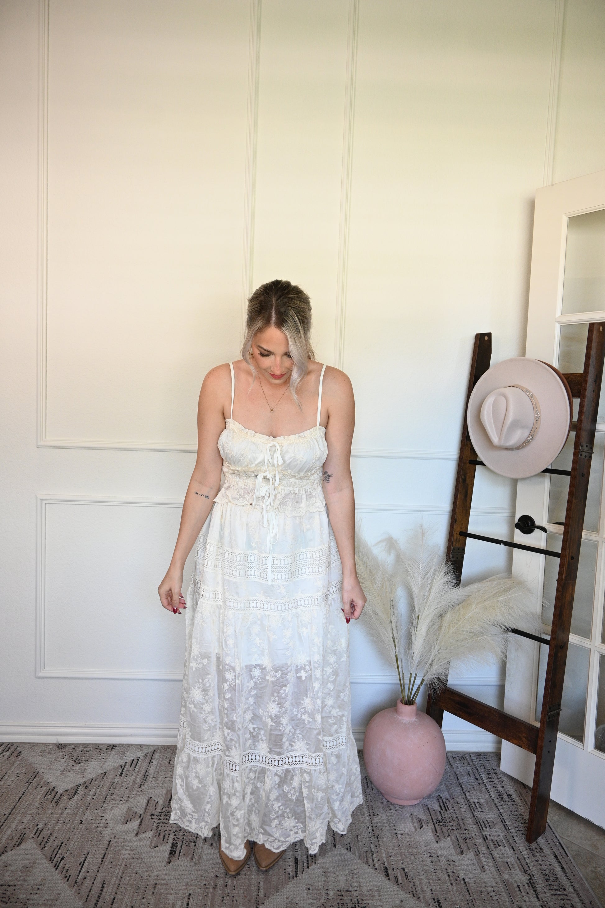 Woman in a ivory tank top and lace skirt set standing next to a hat on a stand in a room with a textured wall.