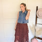 Woman in denim vest and burgundy skirt standing in a room with a white wall and wooden floor.