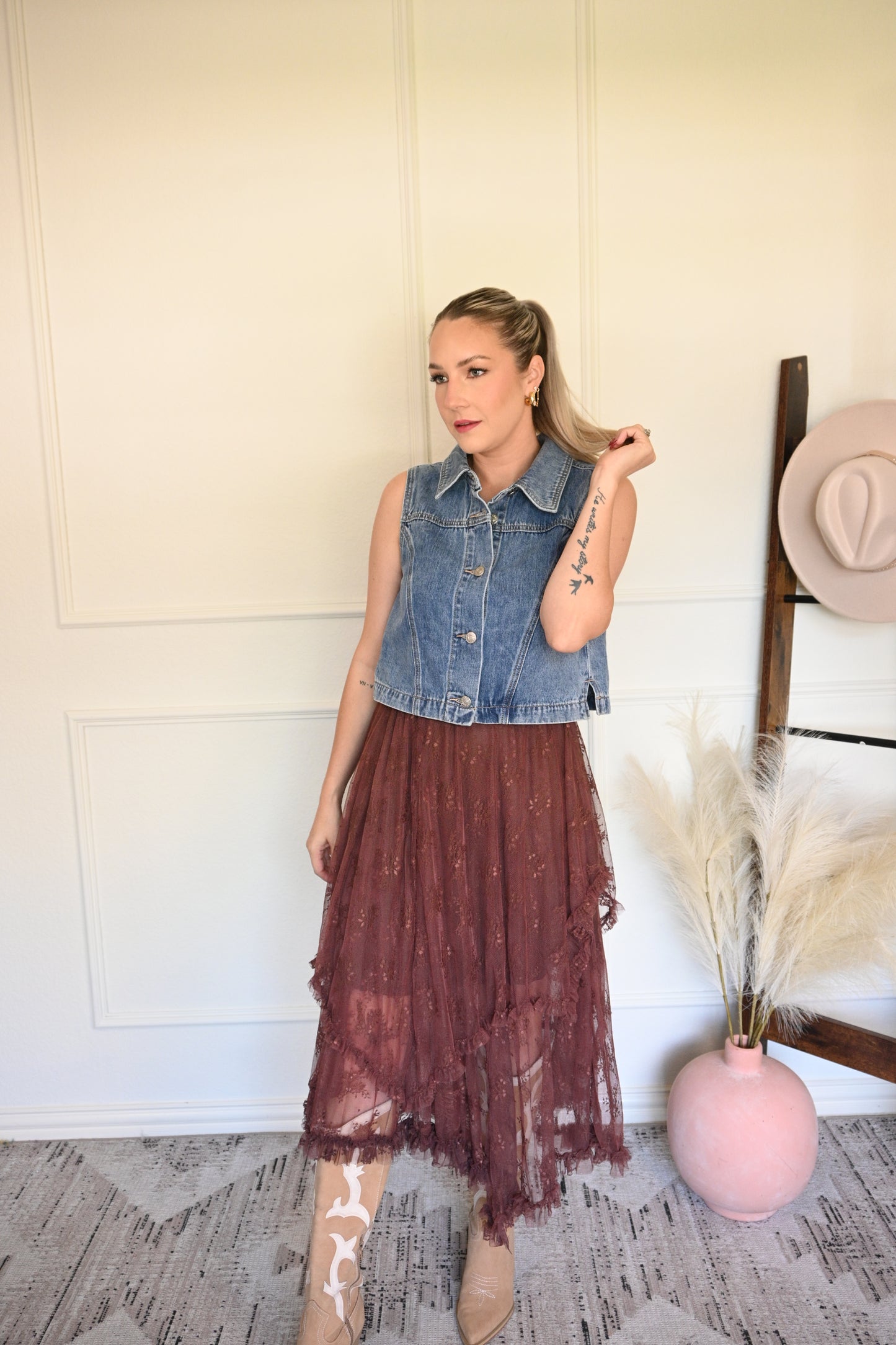 Woman in denim vest and burgundy skirt standing in a room with a white wall and wooden floor.