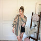 Woman in a striped oversized button down shirt/dress wearing rhinestone black shorts, standing next to a hat rack with hats on a white wall.
