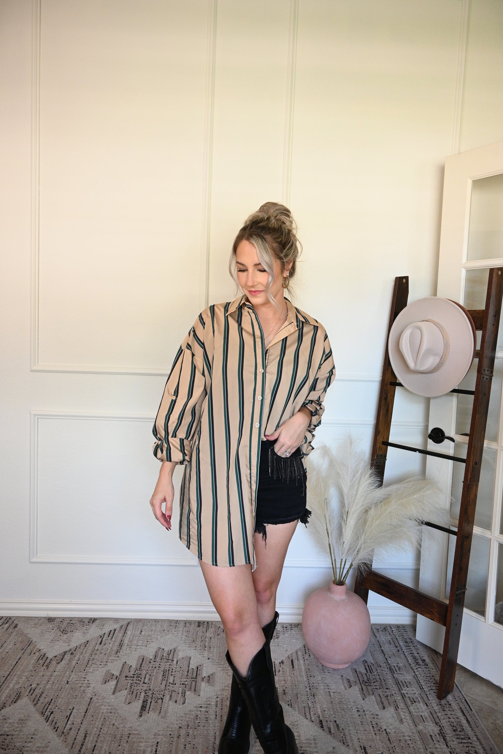 Woman in a striped oversized button down shirt/dress wearing rhinestone black shorts, standing next to a hat rack with hats on a white wall.