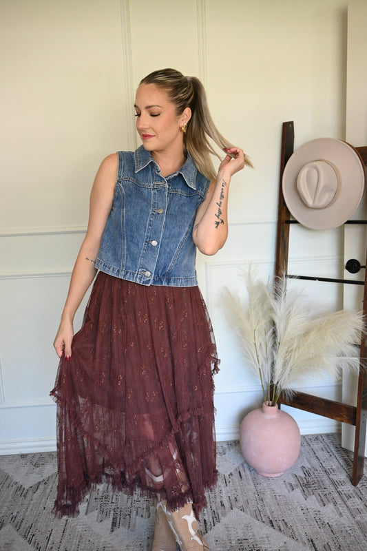 Woman holding her ponytail wearing a denim vest with a brown lace skirt