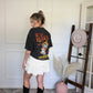 Woman wearing a black t-shirt with text and graphics, cream ruffled shorts, and black boots in a room with a hat rack and decorative items.