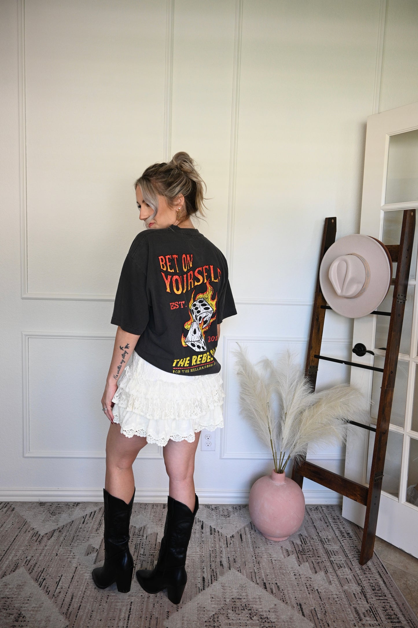 Woman wearing a black t-shirt with text and graphics, cream ruffled shorts, and black boots in a room with a hat rack and decorative items.