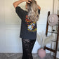 Person wearing a black graphic tee with a graphic design, standing in a room with a white wall and wooden floor.