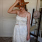 Woman in a ivory lace top and skirt set adjusting a brown hat indoors.
