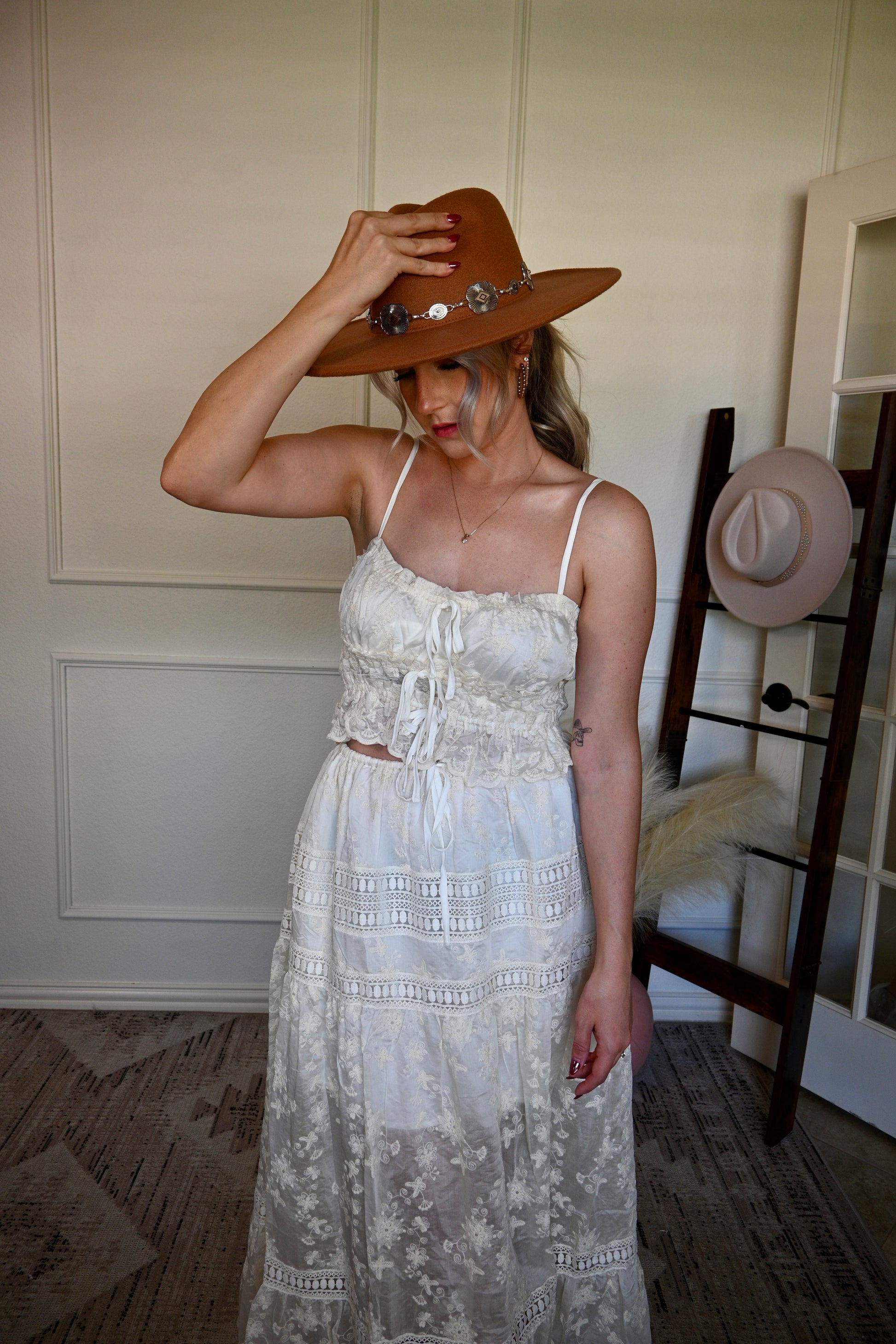 Woman in a ivory lace top and skirt set adjusting a brown hat indoors.