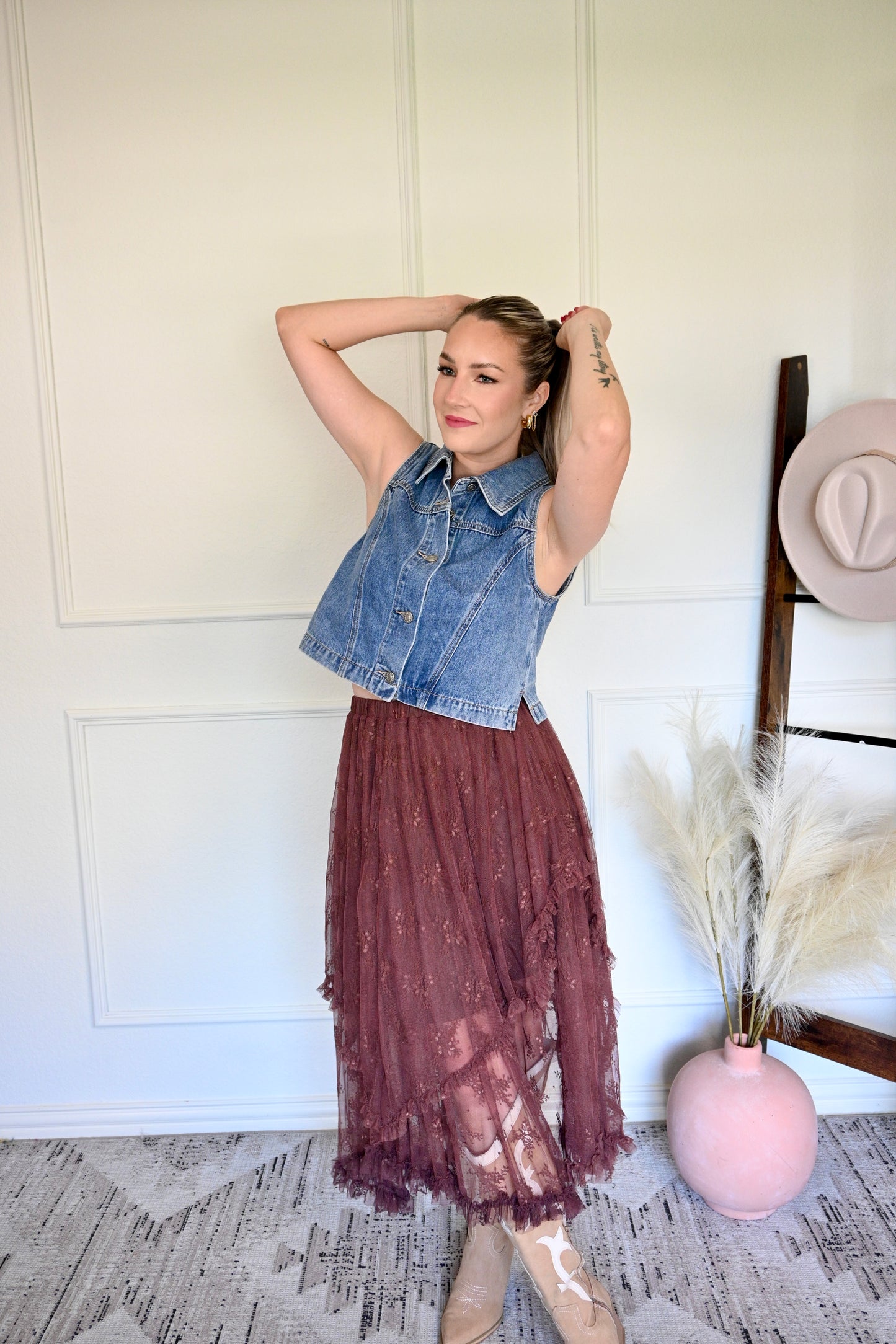 Woman wearing a denim vest over a lace dress in a room with a white wall and decorative elements.