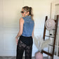 Woman in denim vest and black sheer lace pants standing next to a hat rack with hats on a white wall.