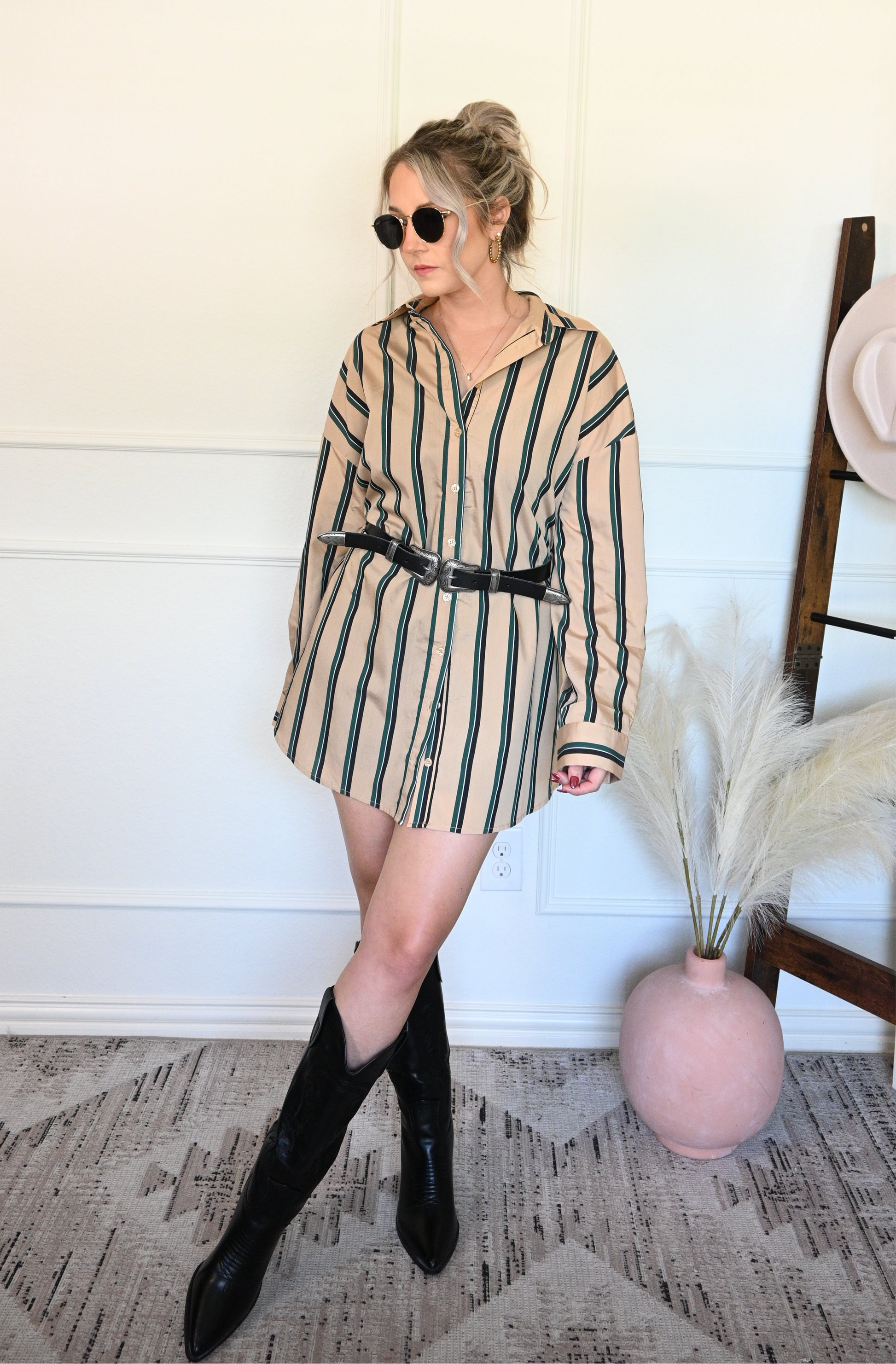 Woman wearing a striped shirt/dress with black boots in a room with a white wall and decorative elements.