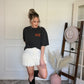 Woman wearing cream ruffled shorts and a black graphic tee in a room with a white wall and patterned rug.
