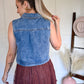 Woman wearing a denim vest with a brown skirt, standing in a room with a white wall and a pink vase.