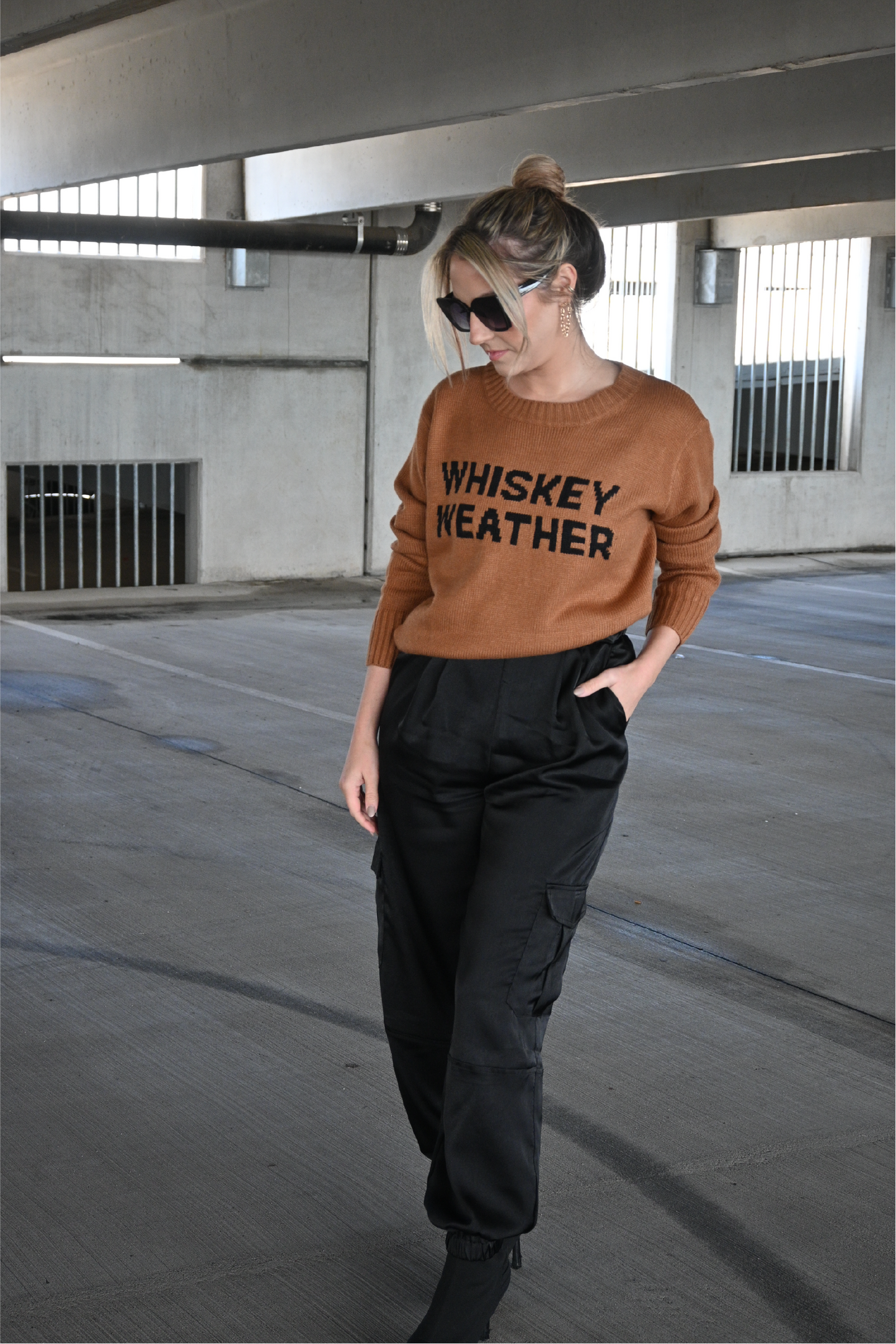 Model is wearing the brown crewneck sweater with the words "Whiskey Weather" in black on the front. It is paired with black satin pants and black heeled boots.