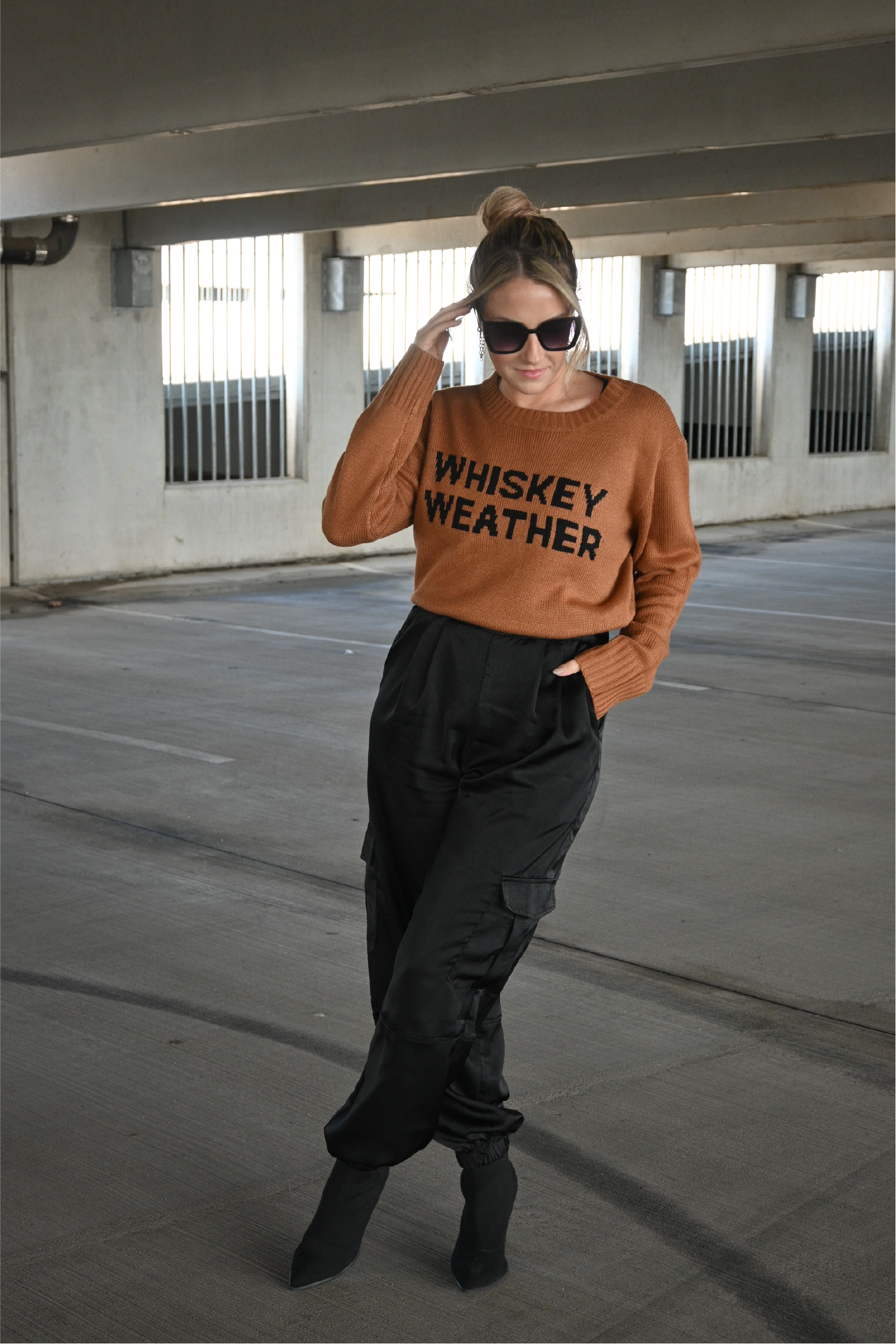 Model is wearing the brown crewneck sweater with the words "Whiskey Weather" in black on the front. It is paired with black satin pants and black heeled boots.