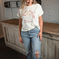 There is a woman standing in front of a white brick wall looking down at the floor. She is wearing light wash high rise skinny jeans and a white vintage graphic tee that says COUNTRY ROADS TAKE ME HOME.