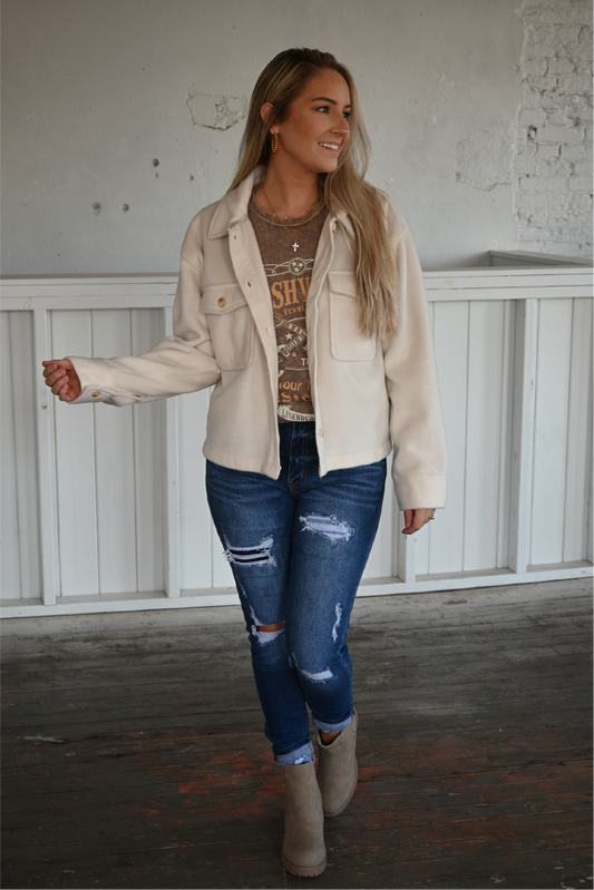 A woman is facing the camera wearing the cropped ecru jacket over the nashville graphic tee paired with the high rise ankle skinny kancan jeans cuffed at the bottom and the sahara slip on booties. 