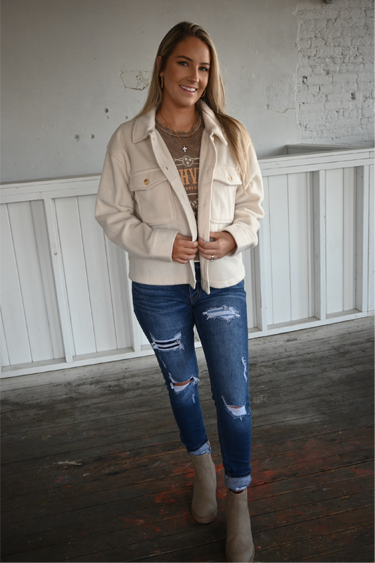 A woman is facing the camera smiling holding the front of the cropped ecru jacket together with the nashville graphic tee underneath paired with the high rise ankle skinny kancan jeans cuffed at the bottom with the sahara slip on booties.