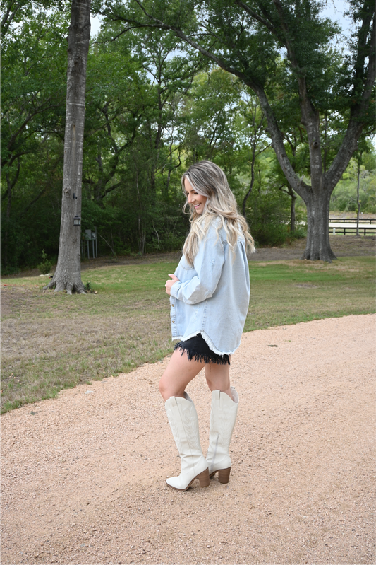 A woman is turned to the side looking down smiling wearing the distressed denim jacket paired with the risen high rise black frayed shorts and ivory knee high MIA boots