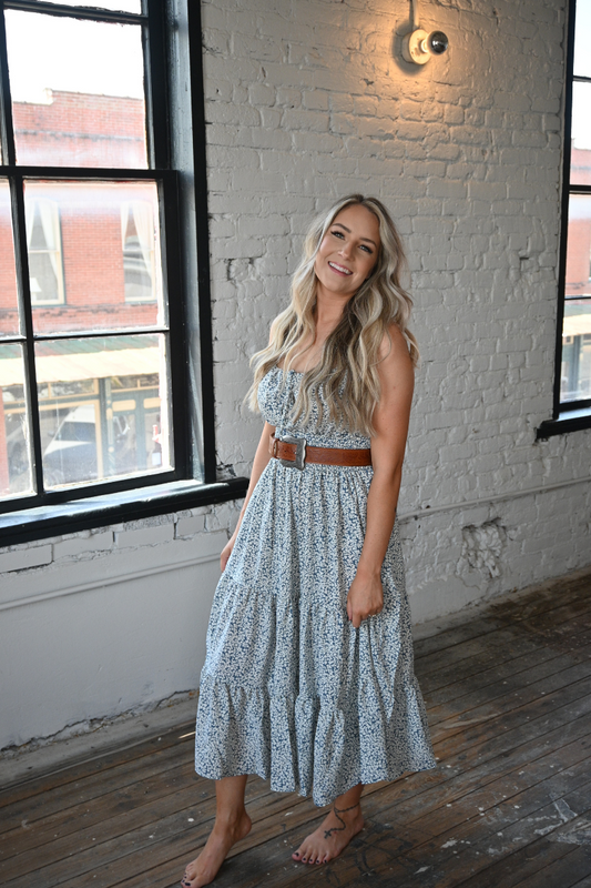 A woman is standing barefoot in front of a white brick wall. She is wearing a blue and ivory floral midi dress.