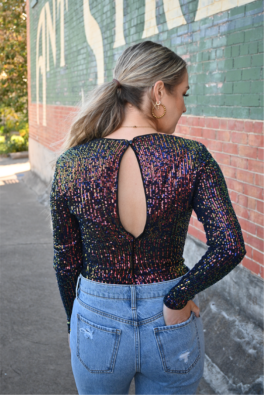 A blonde woman is showing the back of her outfit, she is looking over to her side, with one hand in her back jean pocket. She is wearing a multi color long sleeve sequin body suit. The back of the bodysuit has a keyhole opening with a button closure. She is also wearing our gold beaded earrings.