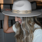 A side view of a blonde woman holding her ivory faux leather band fedora hat.