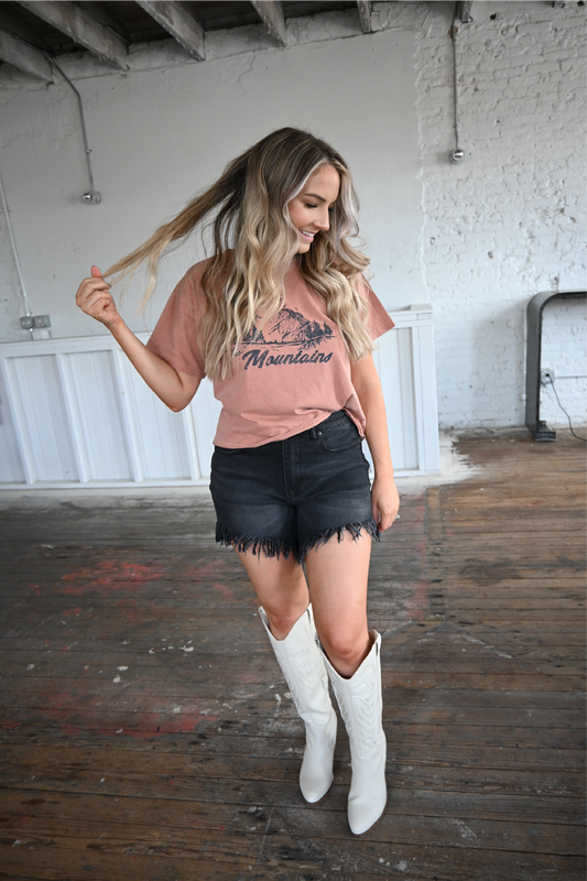 A woman looking to the side smiling with one hand by her side and the other touching her hair wearing a rust colored "take me to the mountains" tee paired with high rise black frayed shorts and ivory knee high boots.