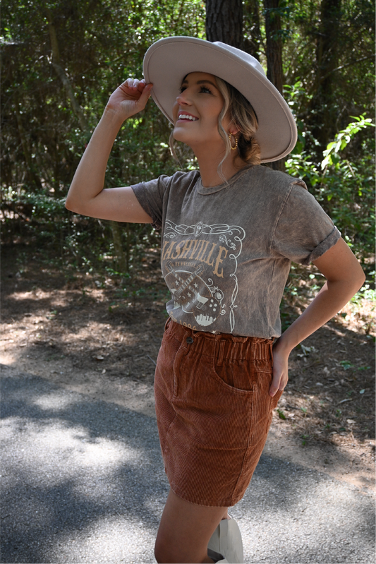 A woman is looking up smiling with one hand on the ivory faux leather band fedora hat and the other hand in her back pocket wearing the nashville graphic tee tucked into the washed corduroy paper bag skirt.