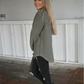 A woman is turned to the side looking at the camera smiling wearing the olive shacket paired with the dark grey skinny jeans cuffed at the bottom with the pierre dumas buckle ankle boots.
