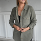 A woman is facing the camera looking off smiling with her hands holding the front of the olive shacket with the sleeves cuffed. she is also wearing the black ribbed cropped cami underneath the shacket paired with the dark grey skinny jeans.