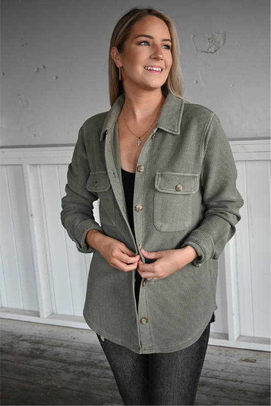 A woman is facing the camera looking off smiling with her hands holding the front of the olive shacket with the sleeves cuffed. she is also wearing the black ribbed cropped cami underneath the shacket paired with the dark grey skinny jeans.