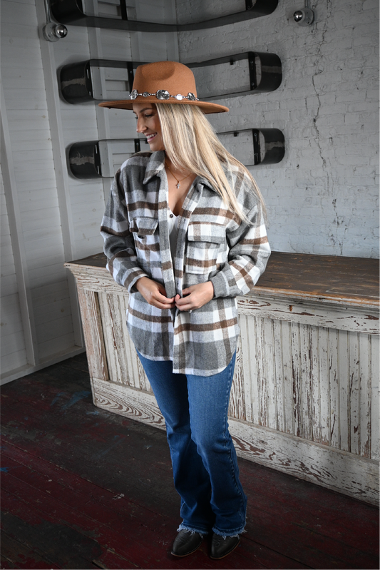 A woman is facing the camera looking down smiling holding the plaid flannel shacket paired with the high rise bootcut kancan jeans, pierre dumas buckle ankle boots and tan silver strap flat brim fedora hat