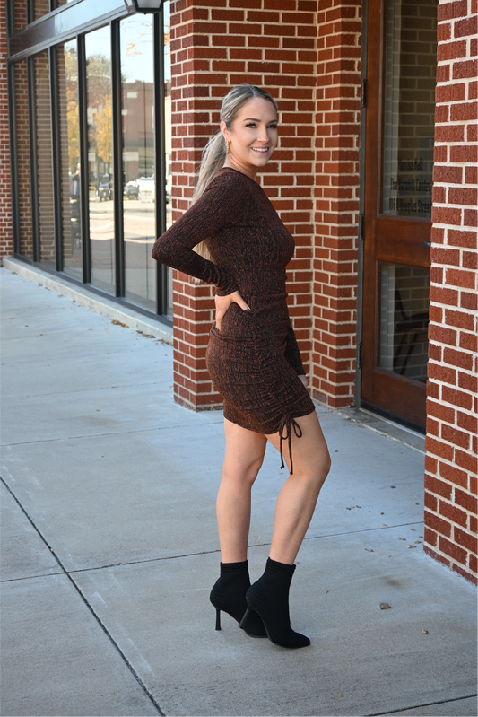 A blonde woman is looking at the camera with the side of her body facing the camera, with one hand on her hip. She is wearing our rust ruched bodycon dress paired with our black fly knit heel boots.