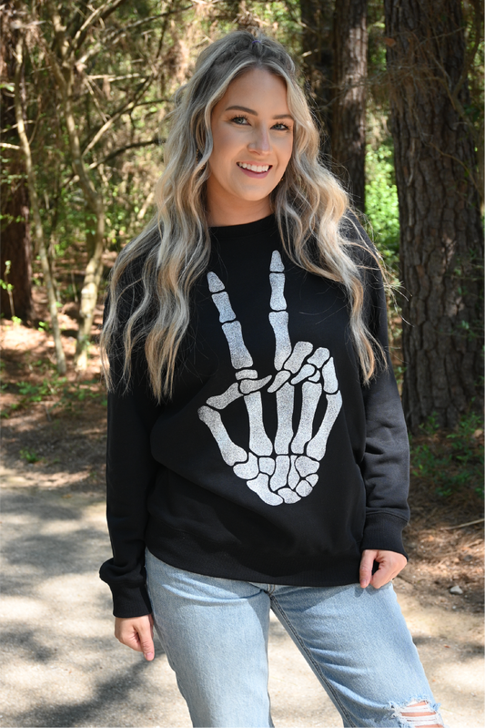 A woman is smiling at the camera wearing the skeleton peace sign sweatshirt paired with the 90's skinny high rise sneak peek jeans.