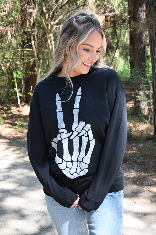 A woman is looking down smiling with her hands in front of her wearing the skeleton peace sign sweatshirt paired with the 90's skinny high rise jeans.