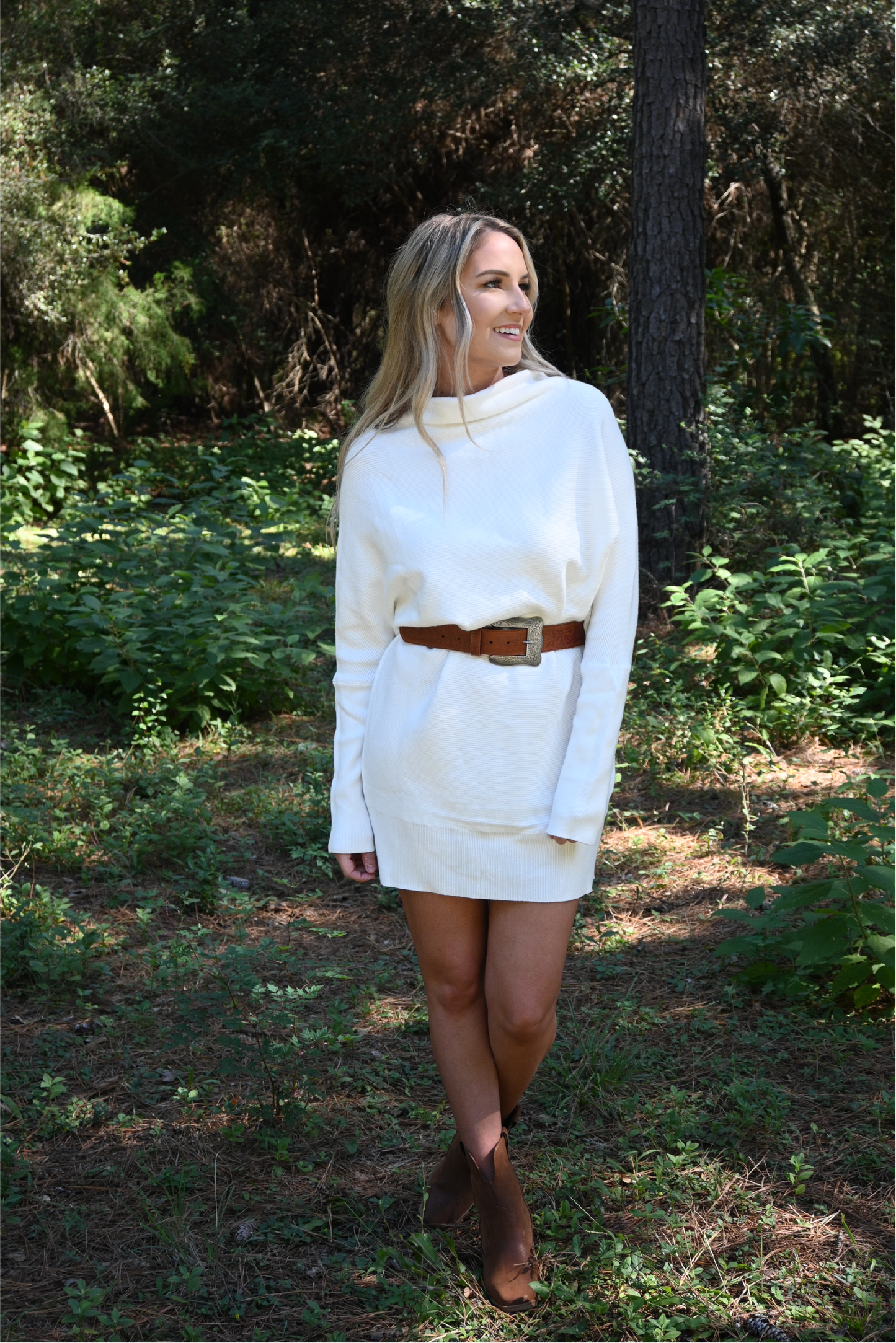 A woman is standing in the woods looking off smiling wearing the slouch neck sweater dress paired with the floral cowgirl buckle belt and western cognac booties.