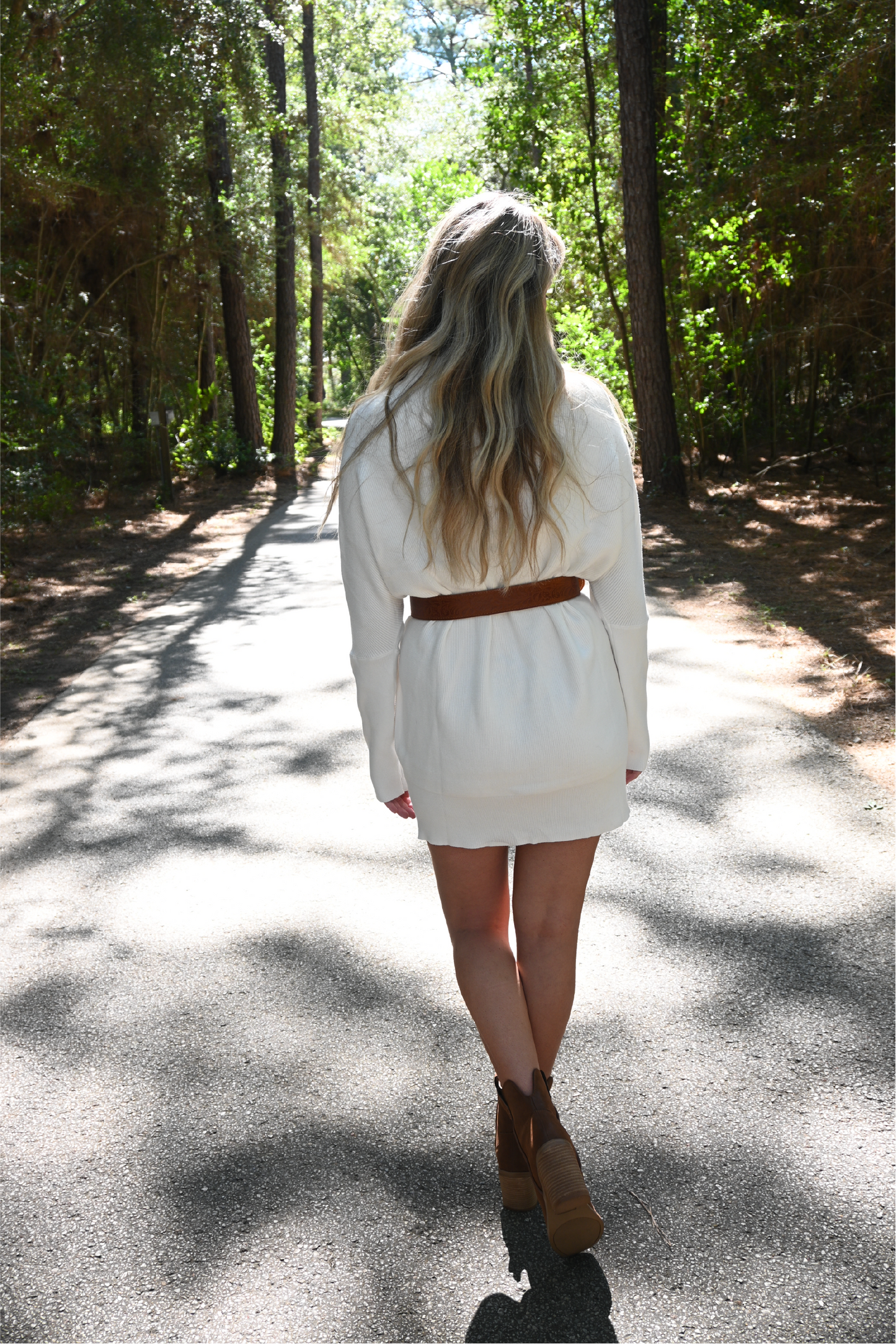 A woman is standing on the road with her back to the camera wearing the slouch neck sweater dress paired with the floral cowgirl buckle belt and western cognac booties.