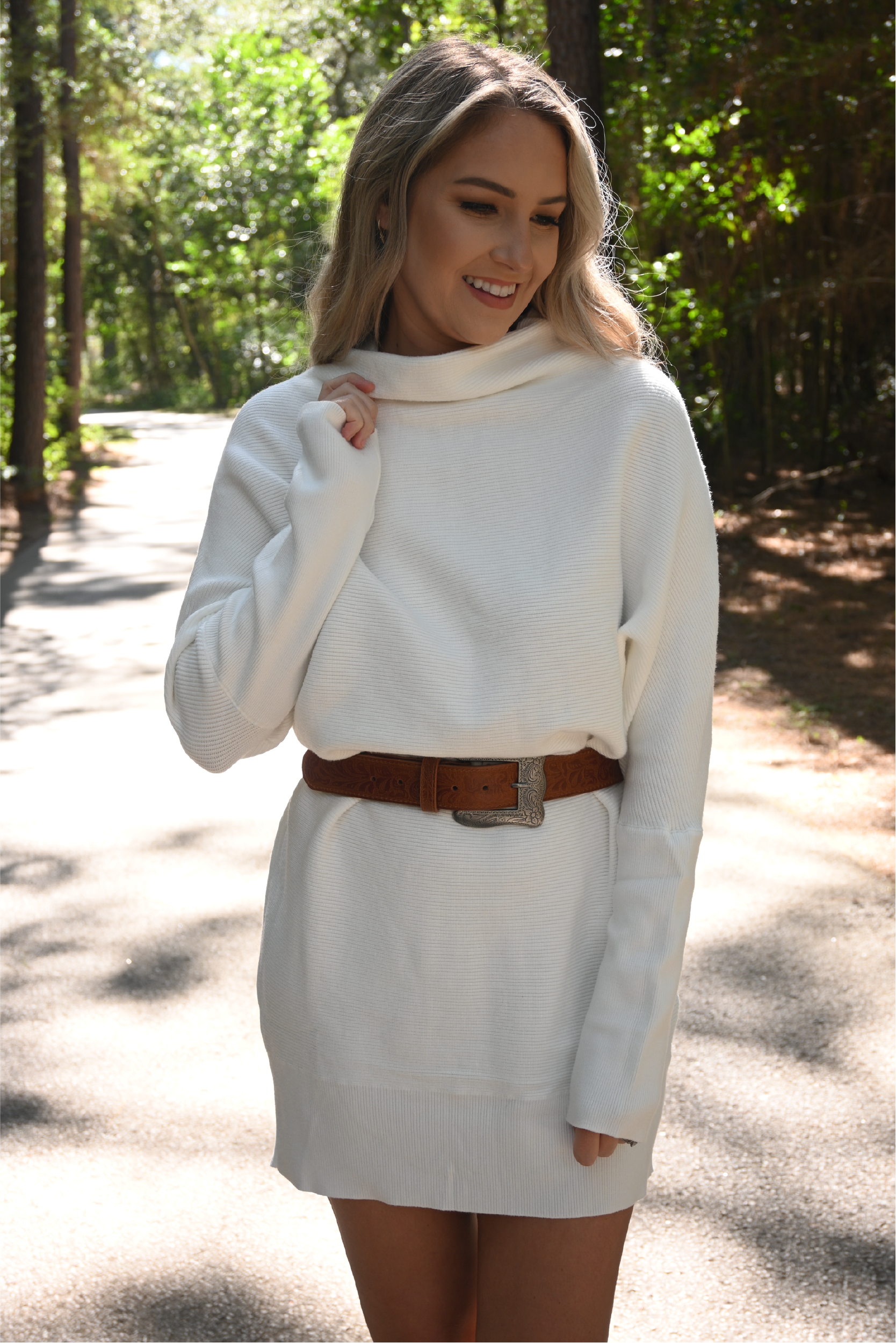 A woman is standing on a road looking down smiling with one hand holding the neckline while the other hand is at her side. she is wearing the slouch neck sweater dress paired with the floral cowgirl buckle belt.