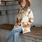 A woman happily sits on a rustic bar wearing an an oversized southwest style jacket, a pair of high rise light wash jeans, a tan fedora hat, and a cream colored graphic tee that says COUNTRY ROADS TAKE ME HOME.