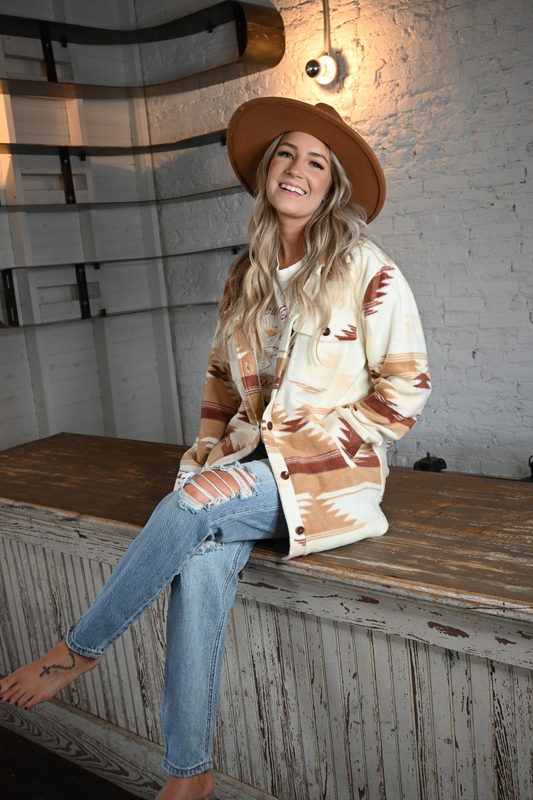 A woman happily sits on a rustic bar wearing an an oversized southwest style jacket, a pair of high rise light wash jeans, a tan fedora hat, and a cream colored graphic tee that says COUNTRY ROADS TAKE ME HOME.