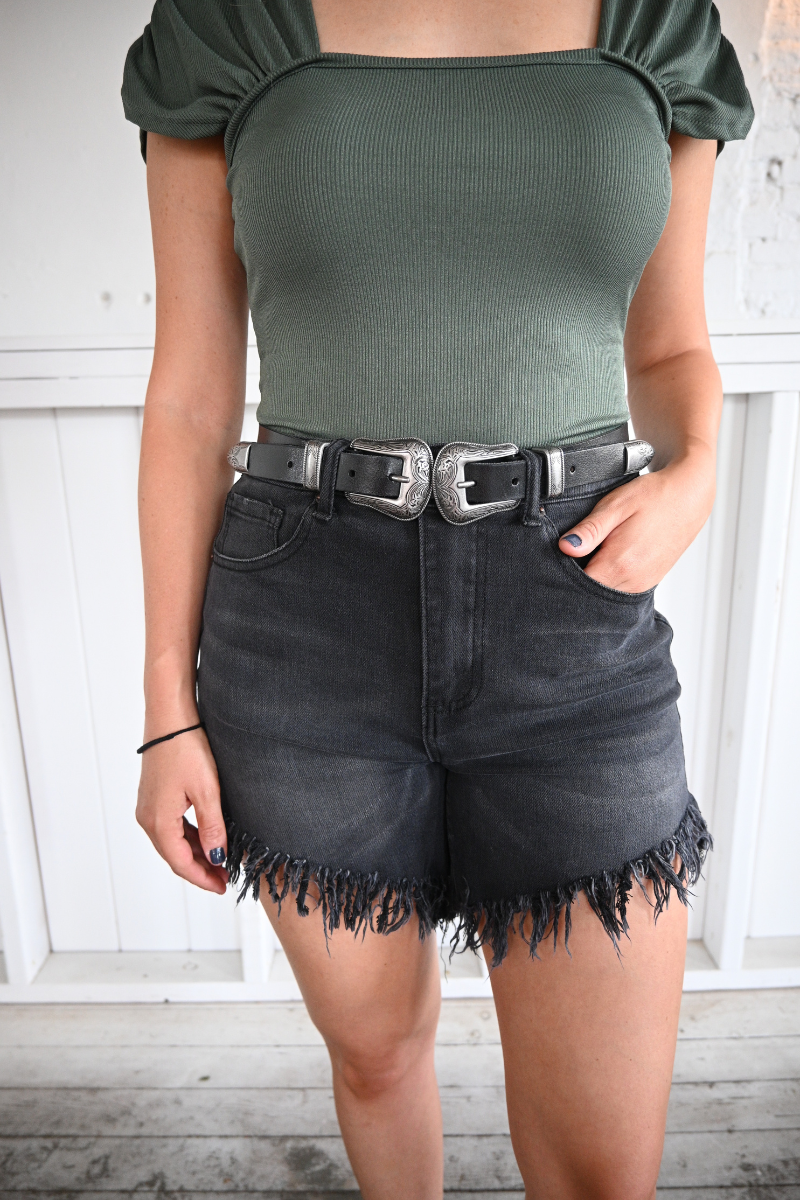 A woman is wearing a hunter green bodysuit and black frayed high rise shorts paired with a black leather belt.