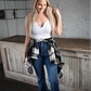 A woman is standing looking down smiling wearing a white ribbed cropped cami with high rise bootcut kancan jeans and a black brown plaid button down around her waist and faded black buckle ankle booties.