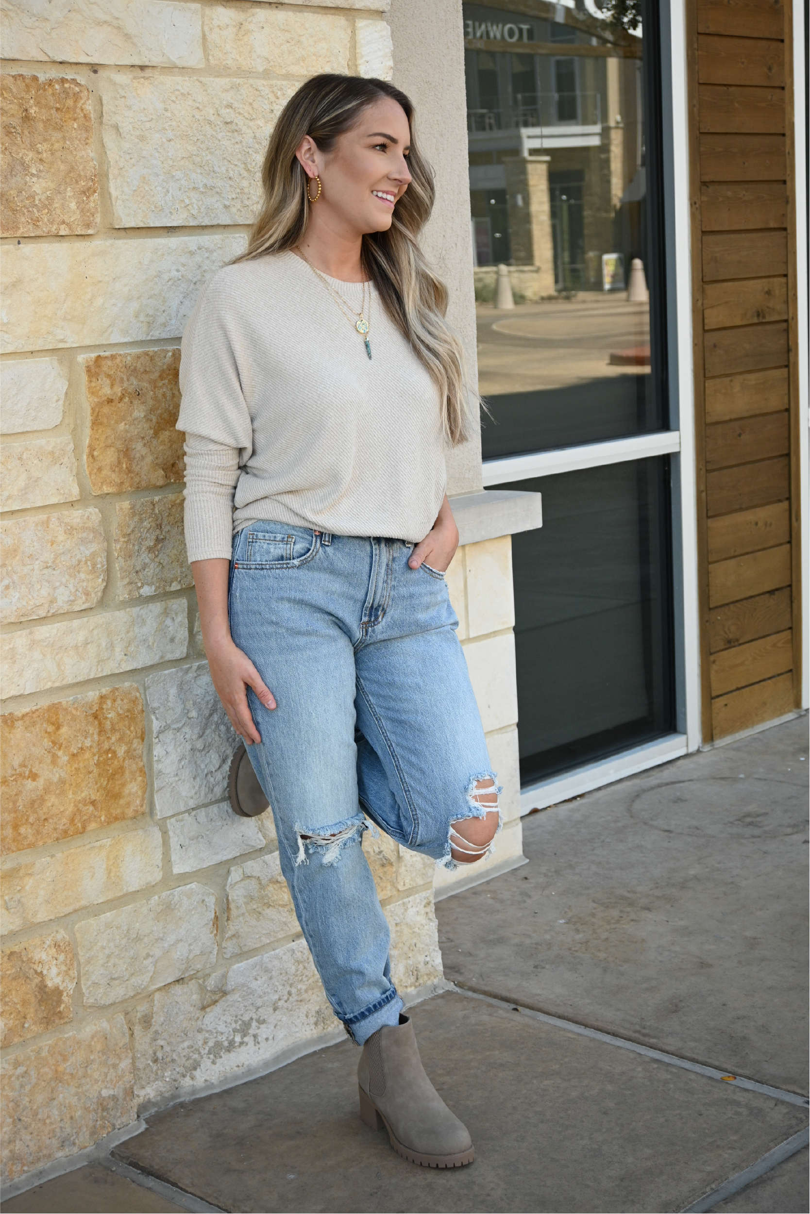 A woman is standing against a stone wall with one foot against the wall while wearing the oatmeal crew neck sweater tucked into the 90's high rise jeans and paired with the sahara slip on booties, also wearing the turquoise spike necklace and turquoise circle necklace