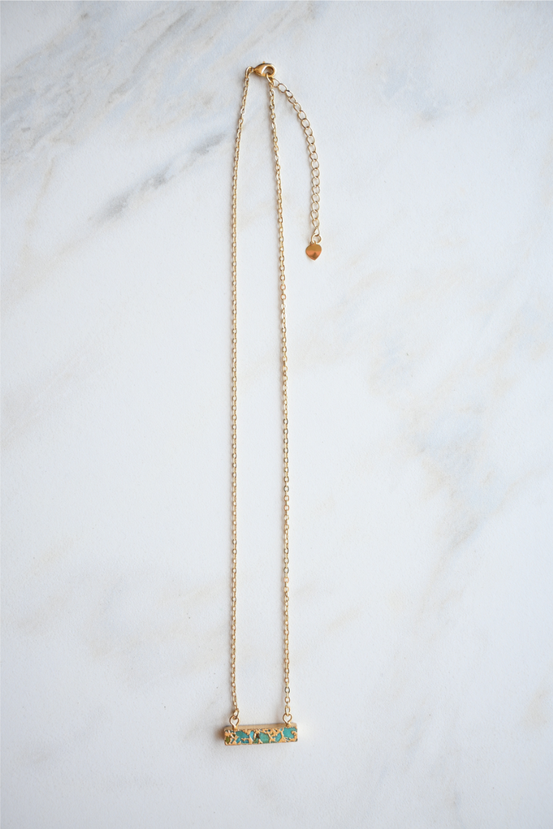 The turquoise bar gold necklace is laying on a white marble tile.