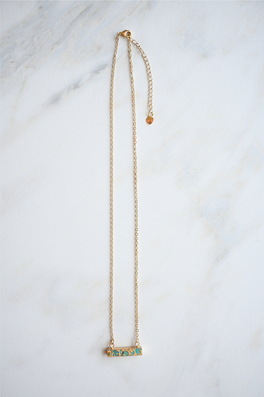 The turquoise bar gold necklace is laying on a white marble tile.